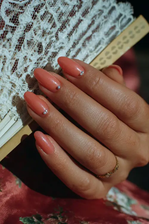 Close-up of bridal hand beside a lace fan, soft coral-pink ballerina nails with glossy finish and dainty silver beads at the cuticle 