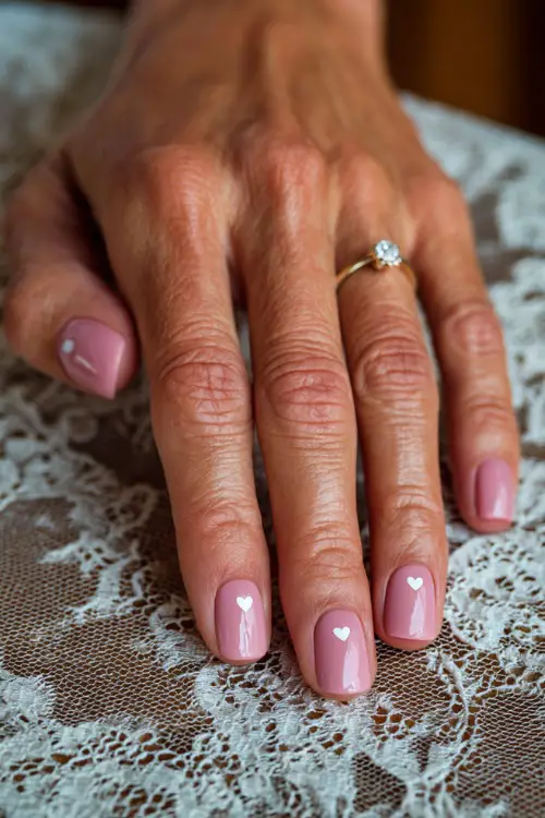 Close-up of bridal hand on vintage lace tablecloth, square-shaped nails in pastel pink with a glossy topcoat and a tiny white heart detail on each ring finger 