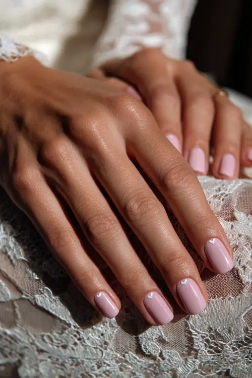 Close-up of bridal hand on vintage lace tablecloth, square-shaped nails in pastel pink with a glossy topcoat and a tiny white heart detail on each ring finger