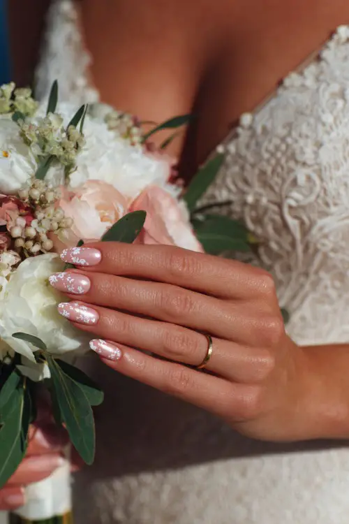 Close-up of bride’s hand holding a summer bouquet, almond-shaped nails in sheer blush pink with glossy finish and delicate white floral decals