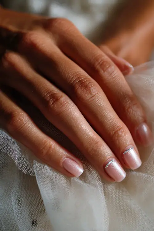 Close-up of bride’s hand on soft white tulle, ballerina nails in creamy pink with chrome shimmer overlay and tiny rhinestones forming a crescent near the cuticle