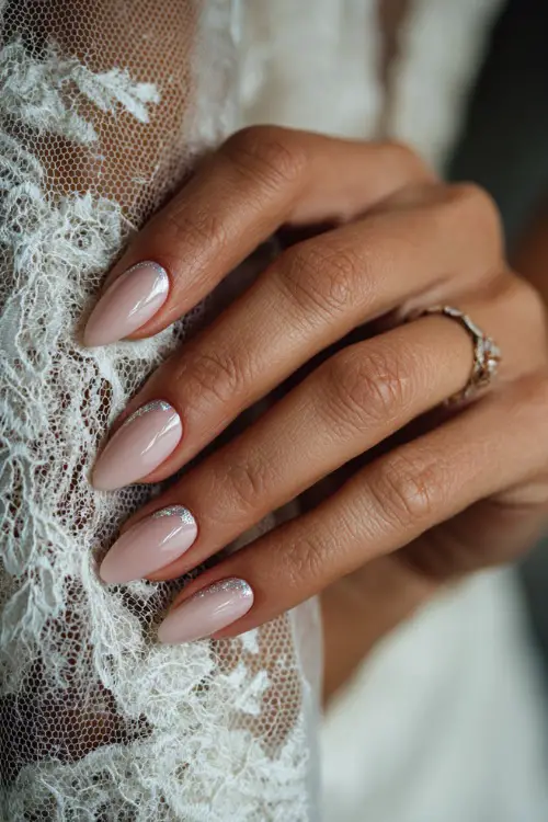 Close-up of bride’s hand resting on a lace veil, almond-shaped nails in soft blush pink with glossy finish and silver foil flakes on two accent fingers