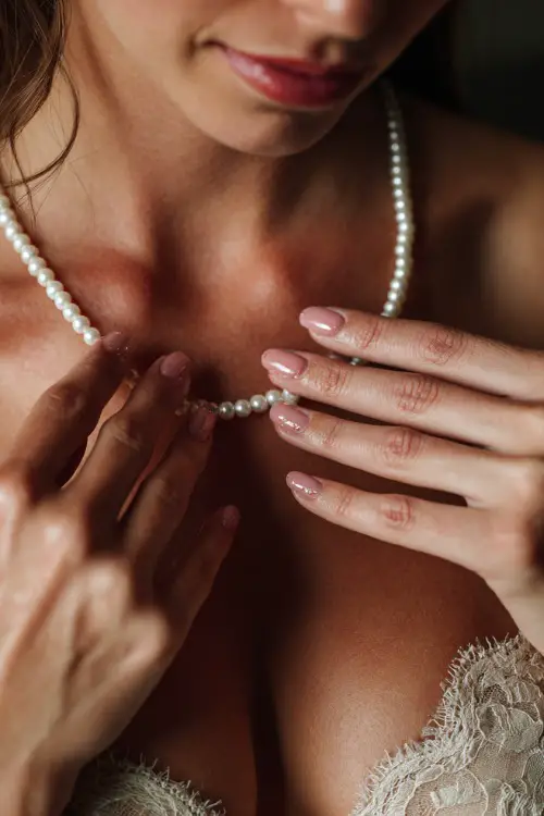 Close-up of bride’s hands gently touching her pearl necklace, nails in light rose pink with tiny pearl studs on ring fingers