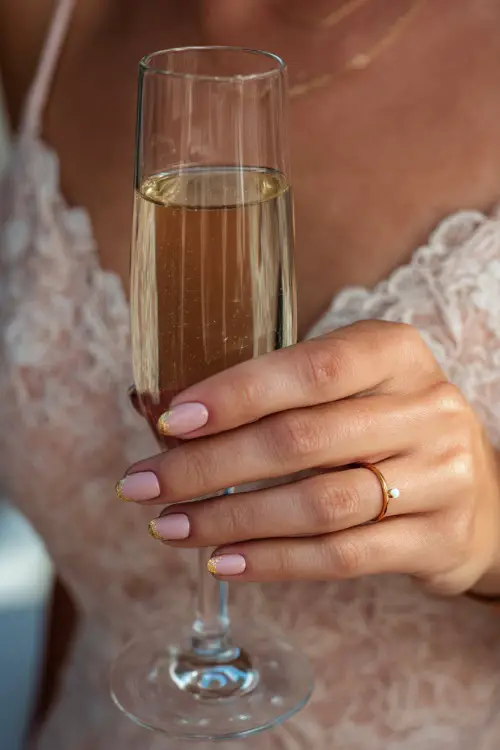Close-up of bridesmaid’s hand holding a champagne glass, short almond nails in soft blush pink with glossy finish and gold micro foil 