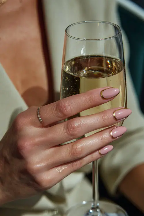 Close-up of bridesmaid’s hand holding a champagne glass, short almond nails in soft blush pink with glossy finish and gold micro foil 