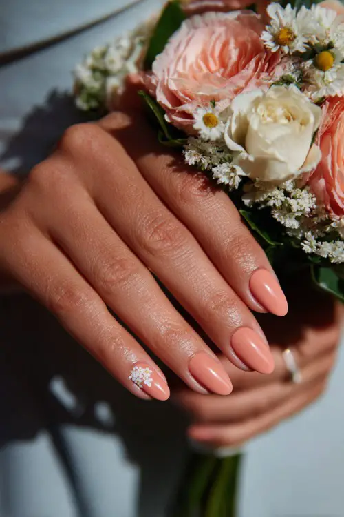 Close-up of bridesmaid’s hand holding a pastel bouquet, short almond nails in soft peach with glossy finish and tiny white floral accents on two fingers 