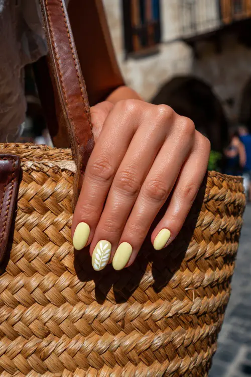 Close-up of guest’s hand beside a woven handbag, short almond nails in pale lemon yellow with glossy topcoat and delicate white leaf design on the middle finger 