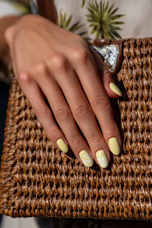 Close-up of guest’s hand beside a woven handbag, short almond nails in pale lemon yellow with glossy topcoat and delicate white leaf design on the middle finger