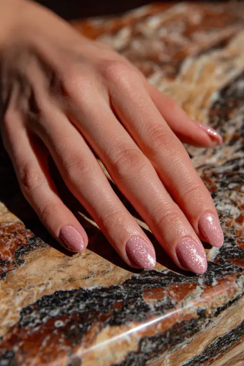 Close-up of guest’s hand on marble table, short oval nails in pastel pink with rose gold glitter dusting across the top half of each nail 
