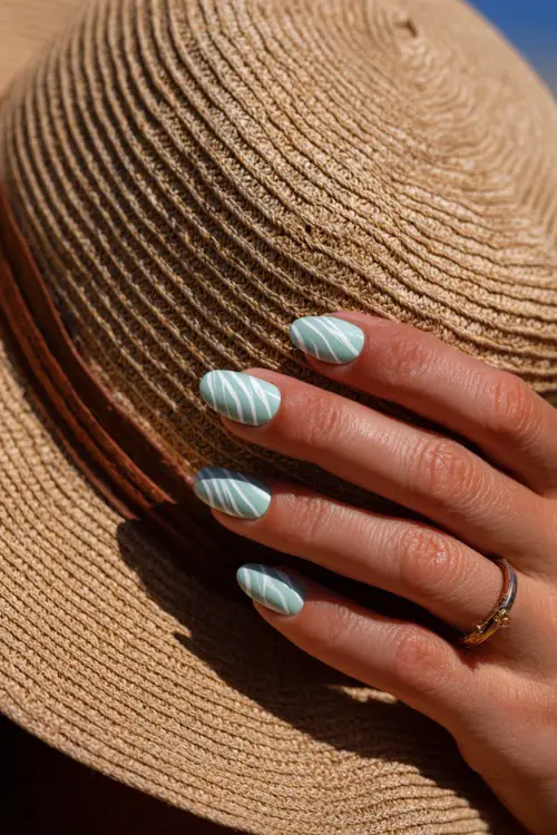 Close-up of hand adjusting a sun hat, almond nails in pastel mint with fine iridescent shimmer and white swirl detailing 