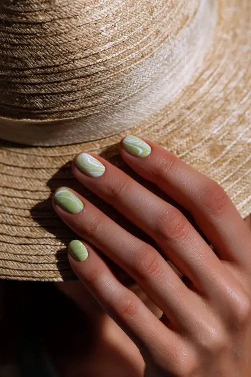 Close-up of hand adjusting straw hat, short ballerina nails in soft lime green with white swirl accent on the ring finger 