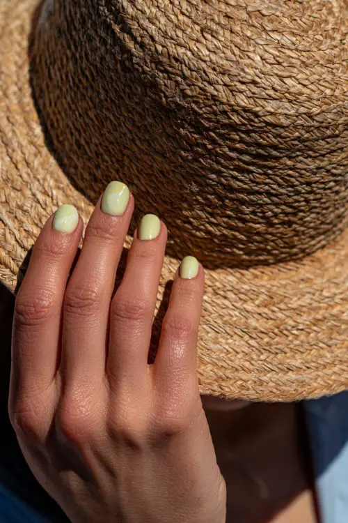 Close-up of hand adjusting straw hat, short ballerina nails in soft lime green with white swirl accent on the ring finger 