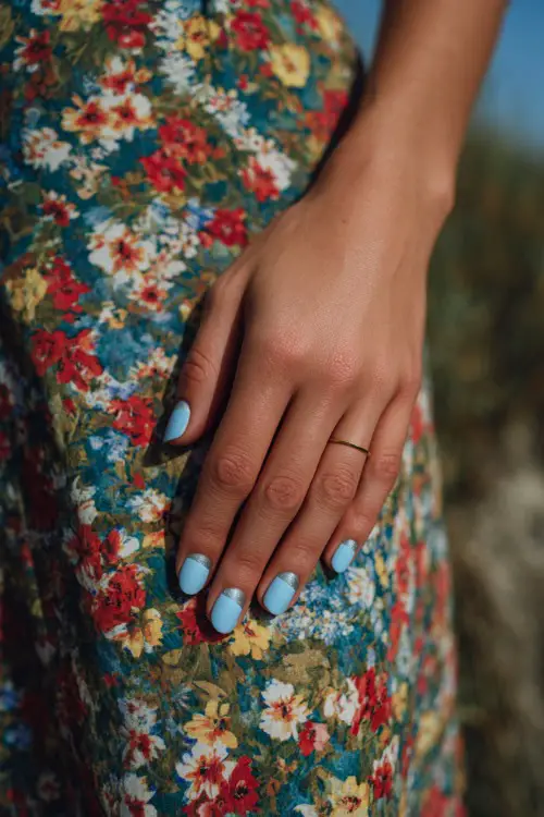 Close-up of hand beside a floral-print bridesmaid dress, short rounded nails in baby blue with silver shimmer on the tips and glossy finish 