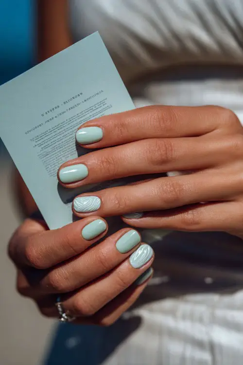 Close-up of hand holding a wedding program, short square nails in pastel mint with one accent nail featuring a white swirl design
