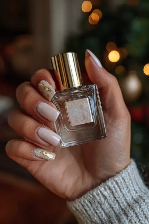 Close-up of hand holding perfume bottle, square-shaped nails in soft beige with marble detailing and gold flake on one nail, romantic lighting in background 