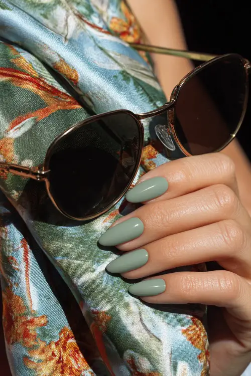 Close-up of hand holding sunglasses, short almond nails in pistachio green with white abstract line art on the ring and middle fingers 