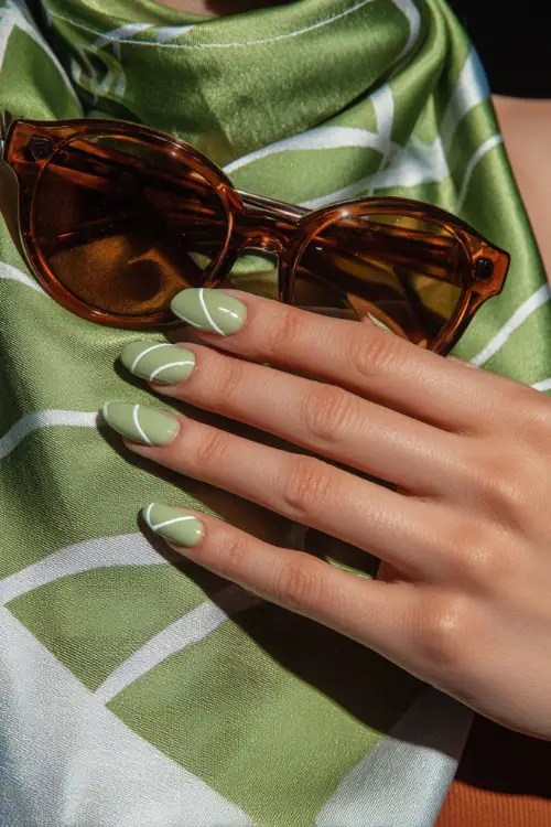 Close-up of hand holding sunglasses, short almond nails in pistachio green with white abstract line art on the ring and middle fingers 