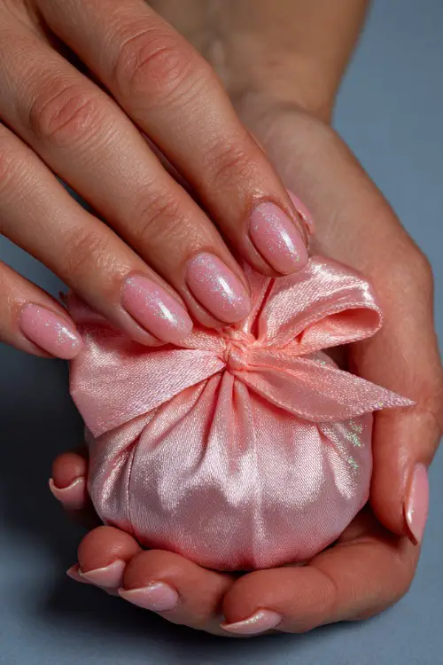 Close-up of hand tying a bow on a wedding favor, short ballerina nails in pastel coral with clear gloss and micro glitter on the pinky finger