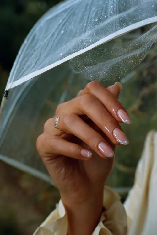 Elegant bride’s hand holding a sheer umbrella, almond nails in translucent rose with silver glitter French tips, outdoor garden wedding light