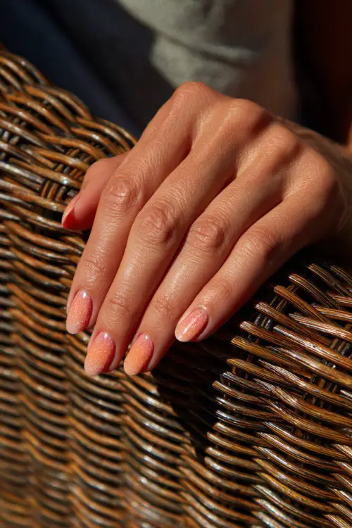 Elegant hand resting on a wicker chair, almond nails in peach ombré with a chrome topcoat and subtle glitter dusting at the tips