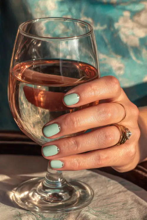 Guest’s fingers holding a glass of rosé, medium square nails in pastel seafoam green with minimal silver dot detail at the base of each nail 