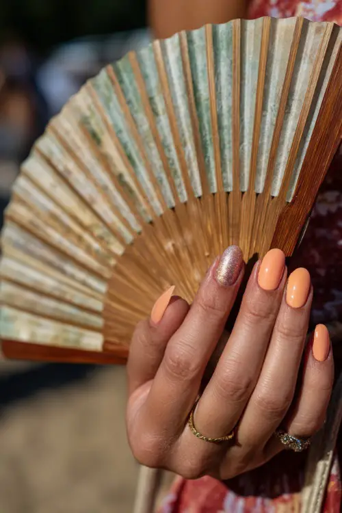 Guest’s fingers holding fan at outdoor ceremony, ballerina nails in peach with a soft ombré fade and one glitter accent nail 
