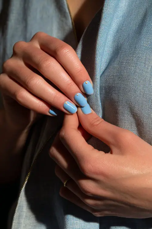Guest’s hand adjusting a linen dress strap, short oval nails in baby blue with glossy finish and minimalist gold dot art near the cuticle