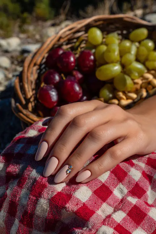 Guest’s hand beside a picnic setup, almond nails in neutral beige-pink with chrome French tips and mini gems on two fingers
