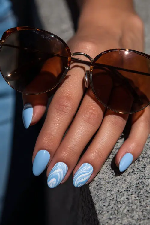 Guest’s hand gently adjusting sunglasses, almond-shaped nails in pastel blue with glossy finish and white swirl design on middle finger