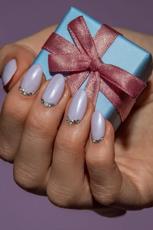 Guest’s hand holding a small ribbon-wrapped gift, short almond nails in pastel lilac with chrome sheen and crystal-studded cuticle design 