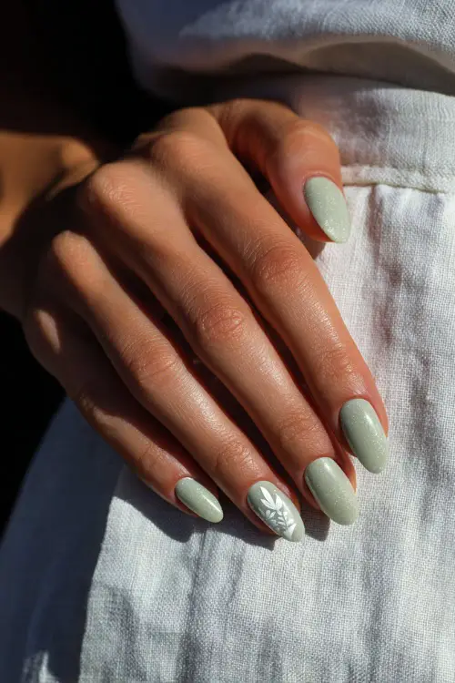 Guest’s hand on white linen, short almond nails in soft sage green with glitter fade and small white leaf decal on the ring finger 