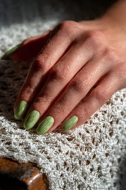 Guest’s hand resting on a white crochet table runner, short nails in pistachio green with marble texture and chrome shimmer 