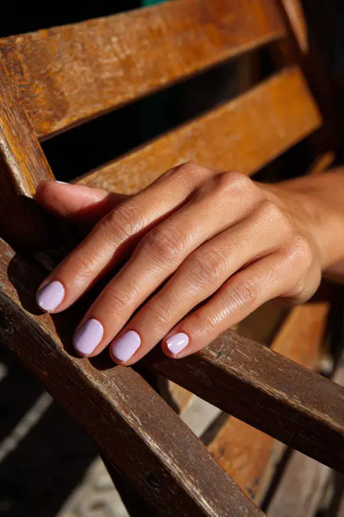 Guest’s hand resting on sun-dappled wooden chair, short nails in pastel lavender with matte finish and single white dot at the tip 