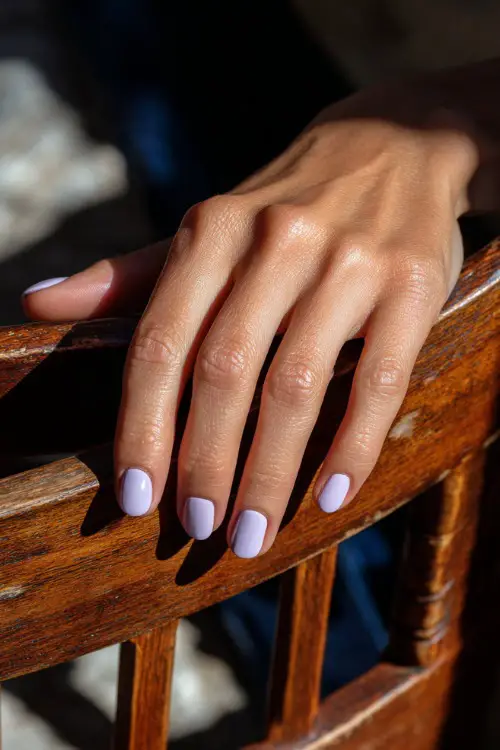 Guest’s hand resting on sun-dappled wooden chair, short nails in pastel lavender with matte finish and single white dot at the tip