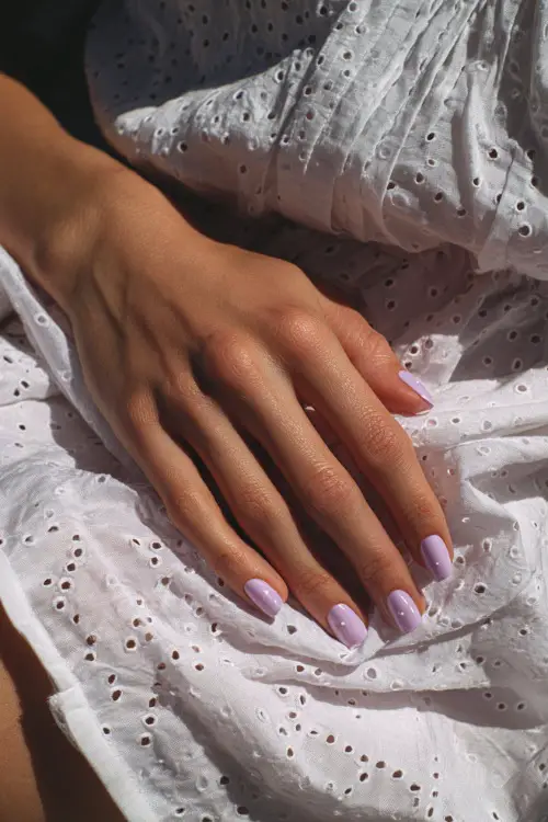 Guest’s hand resting on white eyelet dress fabric, short ballerina nails in pastel lavender with sheer gloss and minimalist dot accents 