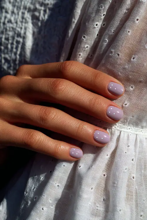 Guest’s hand resting on white eyelet dress fabric, short ballerina nails in pastel lavender with sheer gloss and minimalist dot accents 