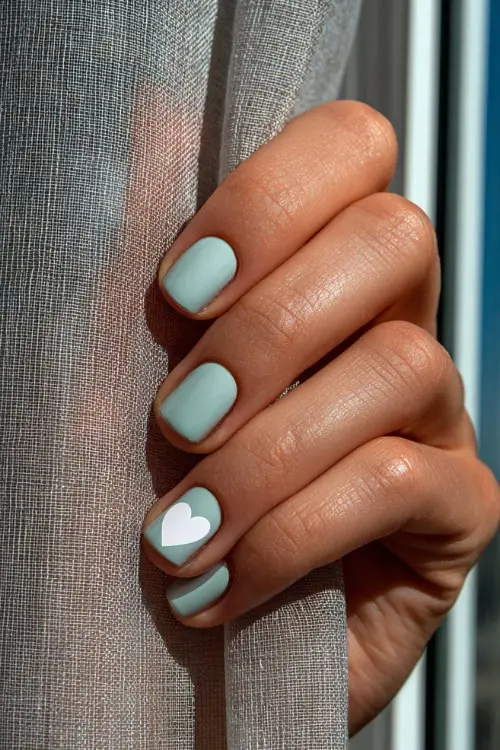 Hand brushing against a sheer curtain, short square nails in pale mint with matte finish and one nail featuring a white heart outline 