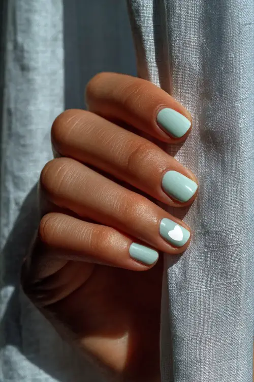 Hand brushing against a sheer curtain, short square nails in pale mint with matte finish and one nail featuring a white heart outline