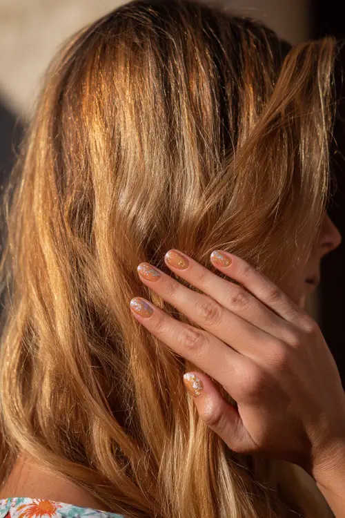 Hand brushing back hair in sunlight, almond nails in soft apricot with gold shimmer topcoat and floral pattern across the tips 