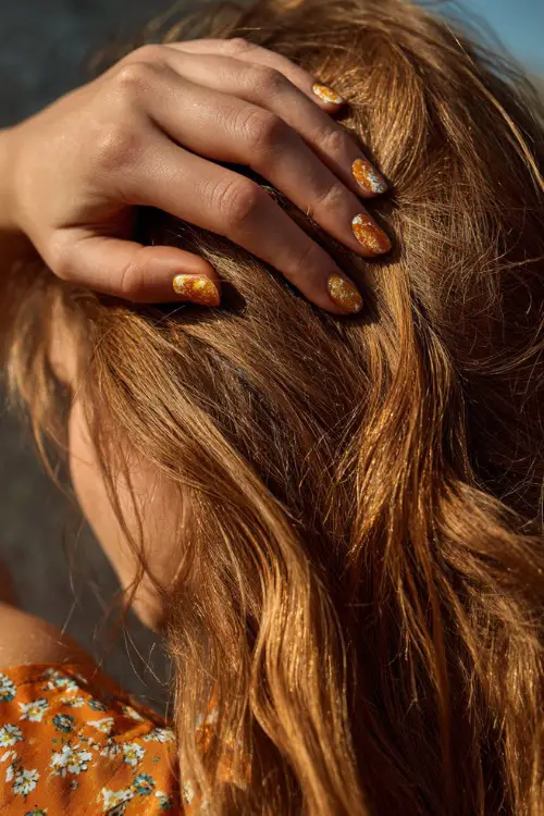 Hand brushing back hair in sunlight, almond nails in soft apricot with gold shimmer topcoat and floral pattern across the tips 