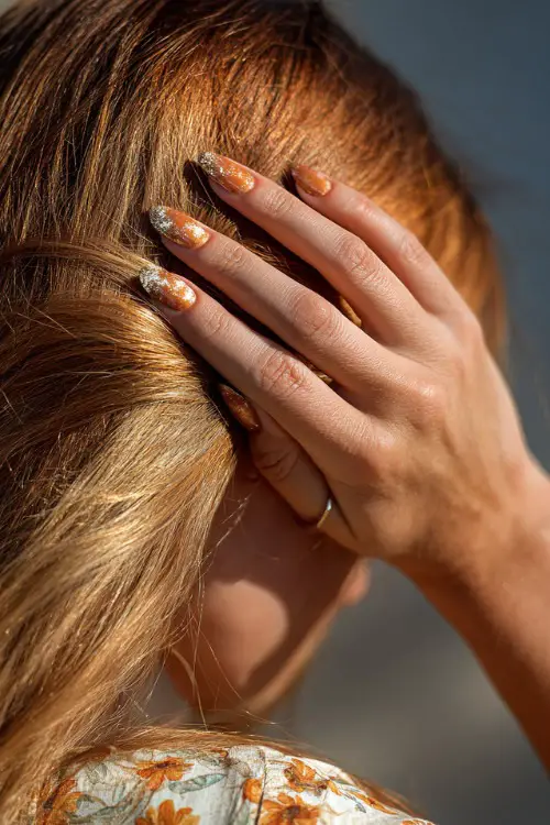 Hand brushing back hair in sunlight, almond nails in soft apricot with gold shimmer topcoat and floral pattern across the tips