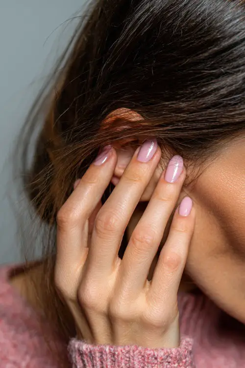 Hand delicately tucking hair behind ear, medium almond nails in soft baby pink with chrome shimmer overlay, dreamy background