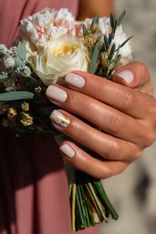 Hand gently holding a bridesmaid bouquet, medium-length square nails in pastel beige with marble accent on one finger and gold foil flakes