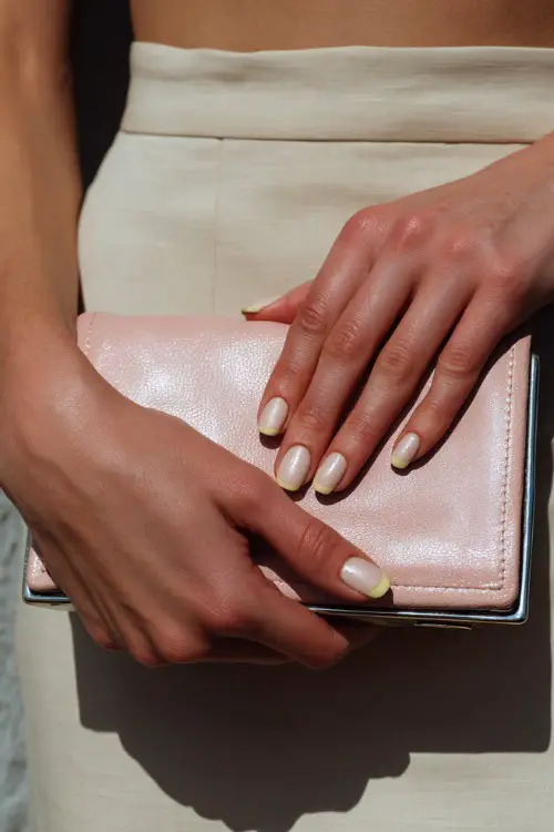 Hand holding a blush pink bridesmaid clutch, square nails in soft ivory with chrome shimmer overlay and thin pastel yellow French tips