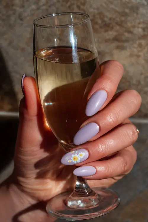 Hand holding a champagne flute, short almond nails in pastel lilac with shimmer overlay and one white floral decal on the thumb 