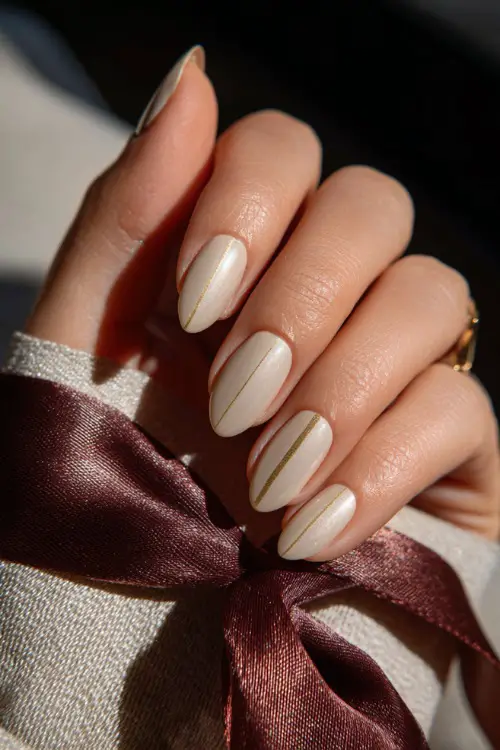 Hand holding a silk ribbon, almond nails in creamy ivory with soft gold foil detailing on two accent fingers, natural sunlight background 