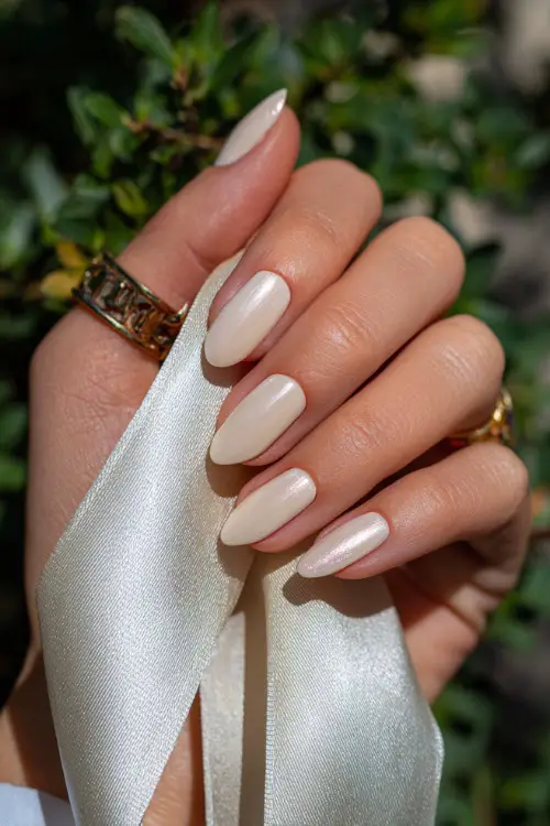 Hand holding a silk ribbon, almond nails in creamy ivory with soft gold foil detailing on two accent fingers, natural sunlight background