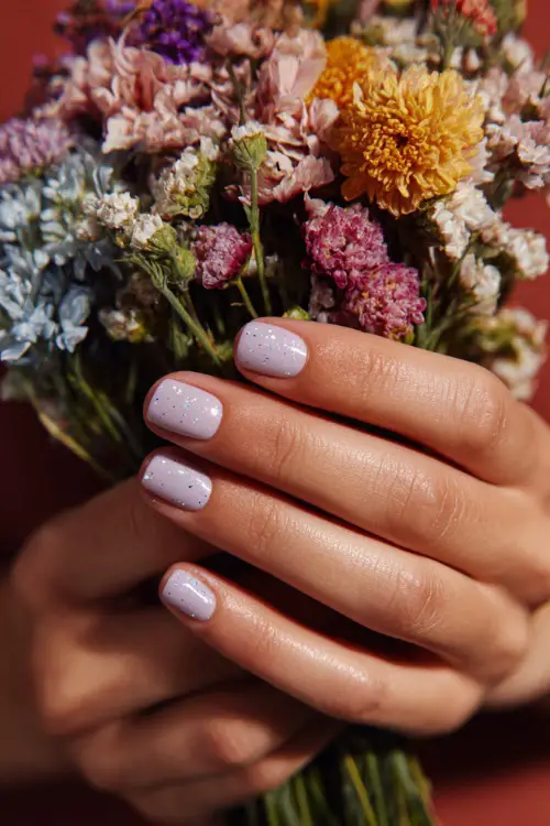Hand holding a summer floral bouquet, short square nails in sheer pastel lilac with tiny silver star flakes