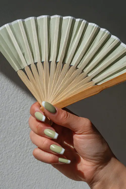 Hand holding a wedding fan, short square nails in pastel olive green with white French tips and shimmer finish