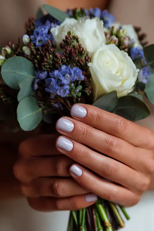 Hand of bridesmaid holding a floral bouquet, square-shaped milky white nails with tiny lavender dot accents on each nail 